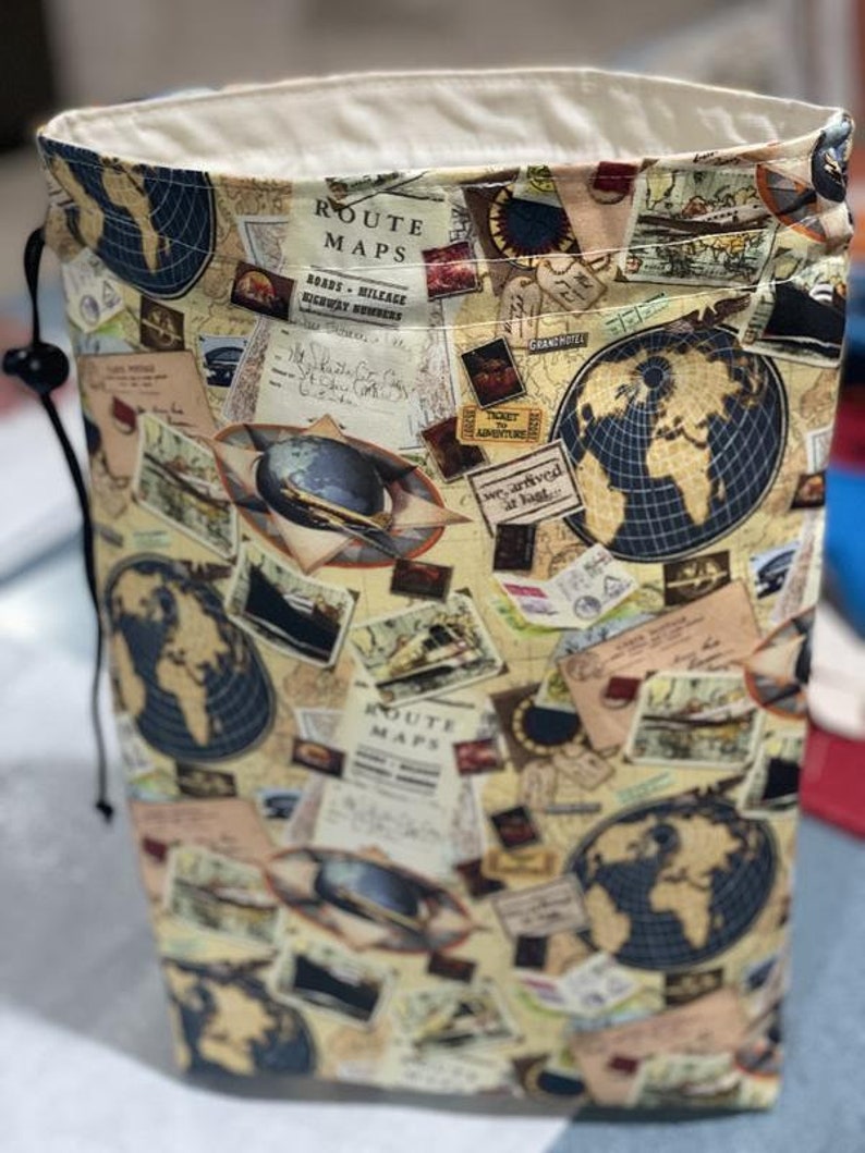 May include: A beige drawstring bag with a vintage travel theme print. The bag features a repeating pattern of maps, globes, and travel-related imagery. The text "ROUTE MAPS" is visible on the bag.