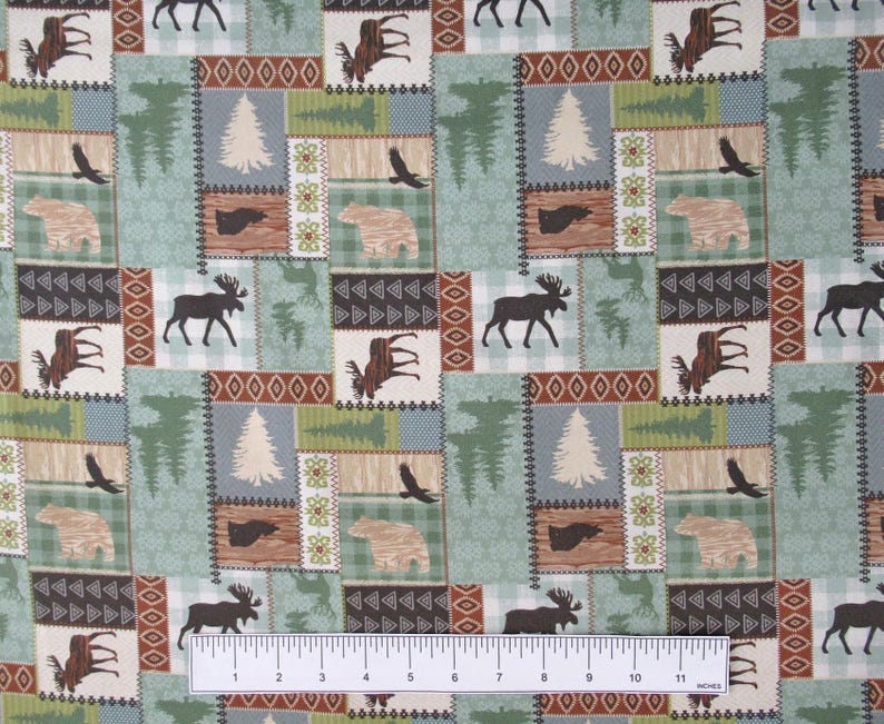 May include: A patchwork fabric with a woodland theme. The design features squares with moose, bears, eagles, and trees in shades of green, brown, and beige. The ruler at the bottom measures up to 11 inches.
