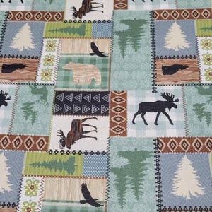 May include: A patchwork fabric featuring woodland animal silhouettes and nature-themed designs. The pattern includes moose, bears, eagles, trees, and geometric shapes in shades of brown, green, and blue. The fabric is likely used for quilting or home decor.