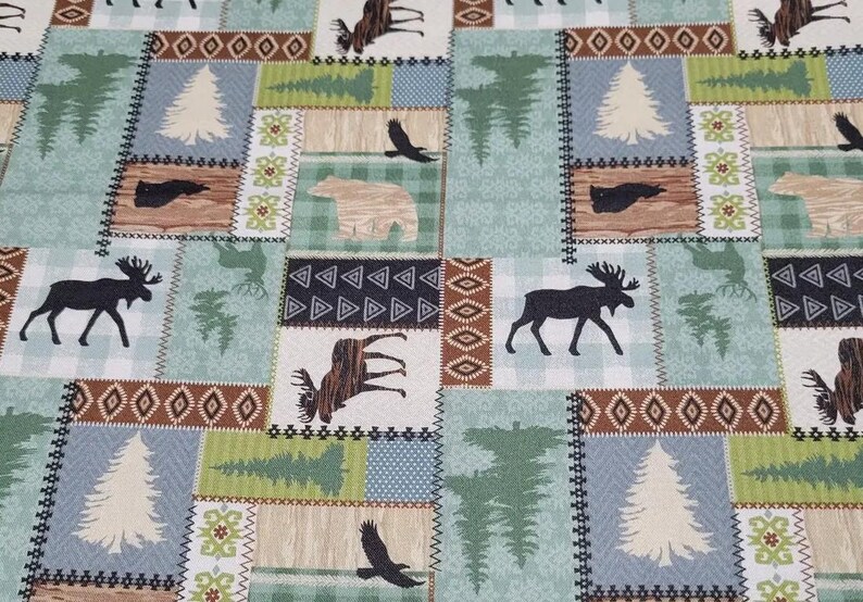 May include: A patchwork fabric featuring woodland animal silhouettes and nature-themed designs. The pattern includes moose, bears, eagles, trees, and geometric shapes in shades of brown, green, and blue. The fabric is likely used for quilting or home decor.