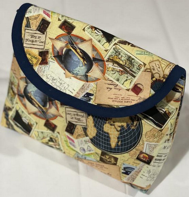 May include: A blue and beige fabric pouch with a flap closure. The pouch is decorated with a vintage travel theme, featuring images of maps, globes, airplanes, and postage stamps. The text "Carte Postale" is visible on the pouch.