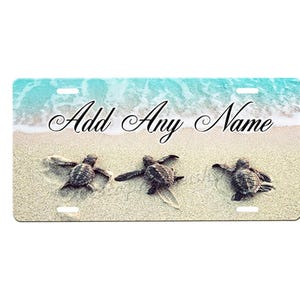 May include: A personalized license plate featuring three baby sea turtles swimming on a sandy beach with blue ocean waves in the background. The text "Add Any Name" is printed on the plate.