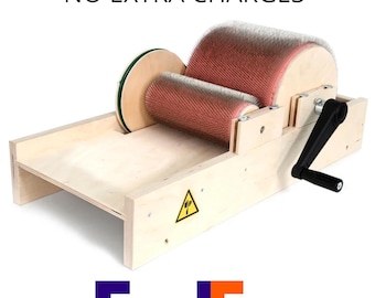 May include: A light-colored wooden drum carder featuring two cylindrical rollers covered in fine metal teeth, a black hand crank on the side, and a yellow triangular warning label with a black icon on the side panel of the wooden frame.