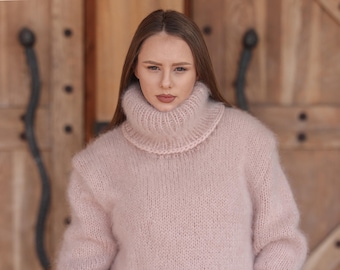Puede incluir: Un jersey de cuello alto rosa claro de gran tamaño. El jersey tiene un cuello alto y acanalado y una textura suave y difusa. Las mangas son largas y el estilo es relajado y cómodo. El fondo es una puerta de madera.