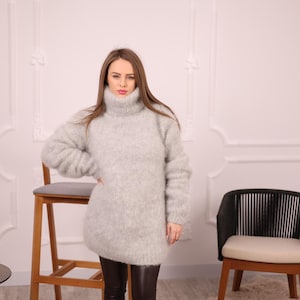 May include: A light gray, oversized turtleneck sweater with long sleeves. The sweater is made of a soft, fuzzy material and is worn with dark brown leather pants. The model is standing in front of a white wall with a chair.
