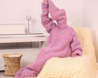 May include: A long-sleeved, hooded sweater dress made of chunky, variegated yarn in shades of pink, blue, and purple. The garment features a thick, loose knit texture, long sleeves, and an integrated hood that extends upward, creating a full-body silhouette.