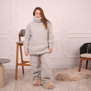 May include: A light gray, fuzzy knit turtleneck sweater and matching pants set. The outfit is worn with light brown fuzzy slippers. The clothing appears soft and comfortable. The model is standing in front of a white wall with decorative molding.