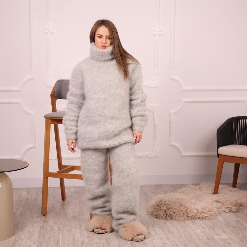 May include: A light gray, fuzzy knit turtleneck sweater and matching pants set. The outfit is worn with light brown fuzzy slippers. The clothing appears soft and comfortable. The model is standing in front of a white wall with decorative molding.