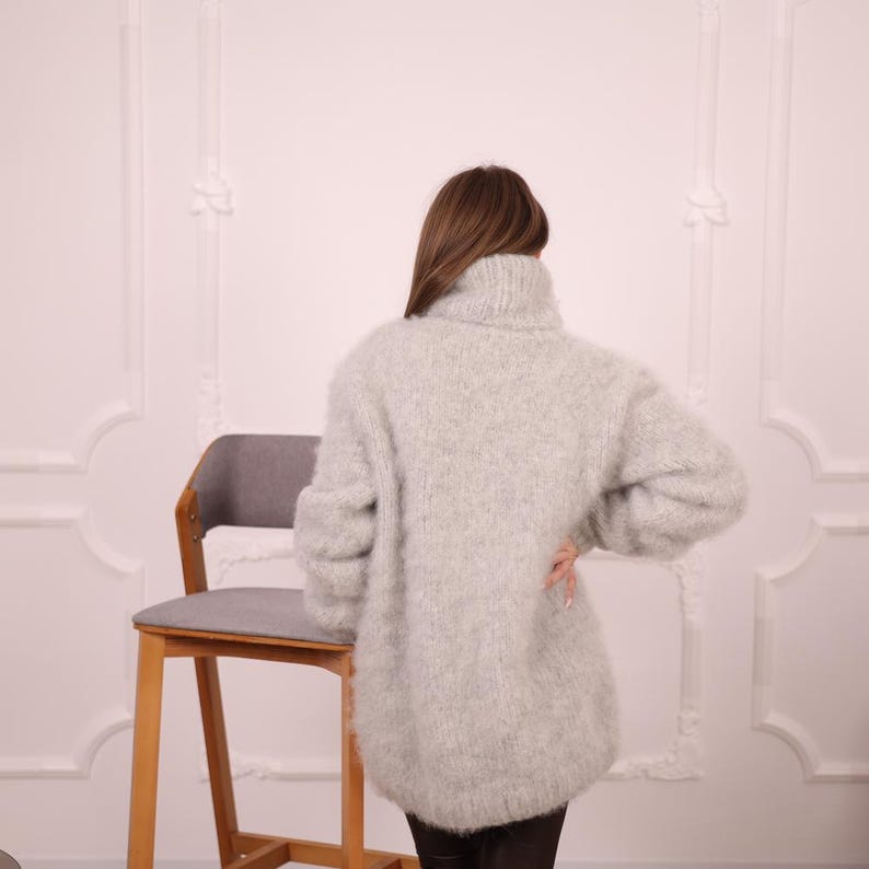 May include: A light gray, oversized turtleneck sweater. The sweater has a loose, comfortable fit and long sleeves. The model is wearing black pants. A wooden chair with a gray seat is in the background.