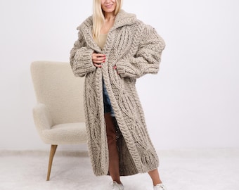 May include: A long, beige, chunky knit cardigan with a shawl collar. The oversized sweater features a cable knit design and is floor-length. The model is wearing the cardigan with a neutral top, denim shorts, and white sneakers.
