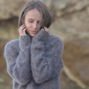 May include: A person wearing a grey, fluffy jumper with long sleeves and a high neck. The jumper looks soft and warm, with a textured appearance. The background is blurred, with a neutral colour palette.