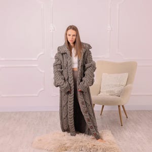 Può includere: Un lungo cappotto grigio a maglia a trecce con chiusura a bottoni sul davanti. Il cappotto ha un ampio colletto e maniche oversize. La modella è in piedi su un soffice tappeto colorato. Il cappotto è un'opzione di abbigliamento esterno comoda ed elegante.