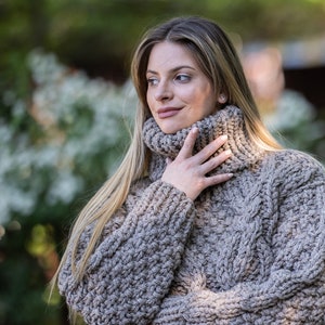 May include: A close-up of a woman wearing a chunky knit turtleneck sweater in a light brown color. The sweater has a cable knit pattern and a high neckline.