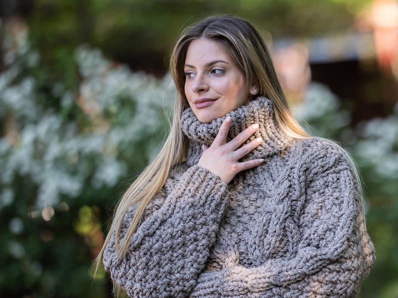 May include: A close-up of a woman wearing a chunky knit turtleneck sweater in a light brown color. The sweater has a cable knit pattern and a high neckline.