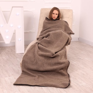May include: A person is wrapped in a large, brown blanket with a black trim, seated in a beige chair. The blanket covers the person from the neck down. A large white letter "M" with illuminated lights is in the background.
