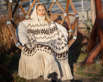 May include: A woman wearing a white and brown knitted hooded poncho with fringe. She is sitting in a white chair with a wooden frame.