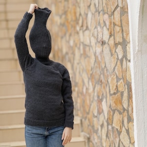 Könnte beinhalten: Ein dunkel anthrazitfarbener, langärmeliger Strickpullover mit einem hohen Rollkragen, der sich nach oben erstreckt und den Kopf bedeckt, kombiniert mit einer blauen Jeans. Der Pullover hat eine gerippte Strickstruktur an Ärmeln und Kragen.