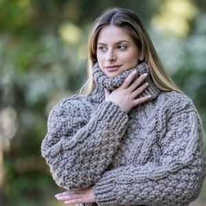 May include: A close-up of a woman wearing a brown chunky knit turtleneck sweater. The sweater has a cable knit pattern and is made of a soft, warm material.