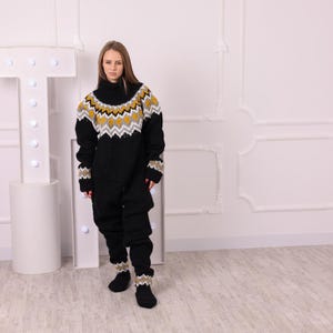 May include: A black knitted jumpsuit with a high neck and long sleeves. The upper part of the jumpsuit features a geometric pattern in yellow, white, and gray. The cuffs and ankles have matching patterned details. The model is wearing black socks.