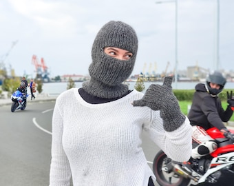 May include: A gray knitted balaclava and matching fingerless gloves. The balaclava covers the head and neck, with openings for the eyes and mouth. The gloves have a ribbed design. The person is wearing a white sweater.