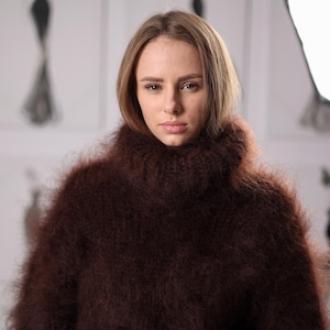Peut inclure: Un pull à manches longues marron foncé doté d'un col roulé haut et épais. Le vêtement est confectionné dans une matière en maille texturée et duveteuse, avec un poil visible, doux et velu qui recouvre toute la surface du pull.