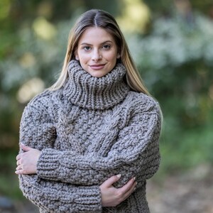 May include: A close-up of a woman wearing a chunky knit turtleneck sweater in a light brown color. The sweater has a cable knit pattern and is made of a soft, warm material.