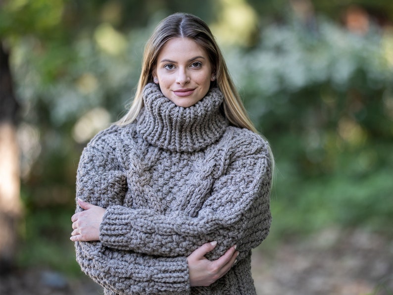 May include: A close-up of a woman wearing a chunky knit turtleneck sweater in a light brown color. The sweater has a cable knit pattern and is made of a soft, warm material.