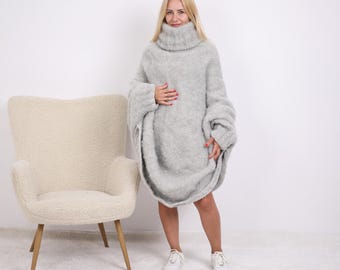 May include: A light grey, oversized turtleneck sweater dress. The dress features long sleeves and a loose, flowing shape. The model is wearing white trainers. The background is a plain white wall and a cream-coloured chair.