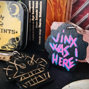 May include: A black wooden keychain with a skull design and the words "Jinx Was Here!" written in pink and blue paint.