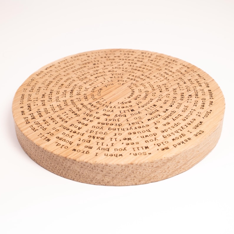 Puede incluir: Un posavasos redondo de madera con texto grabado en espiral. El posavasos es de madera clara y tiene un acabado natural. El texto parece ser un poema o una cita.