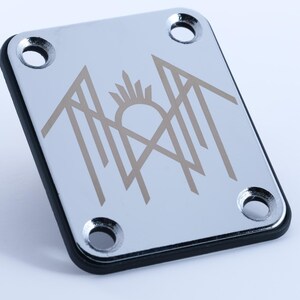 May include: A chrome guitar neck plate with a black border and four screw holes. The plate features a stylized design in a light brown colour, including a sun and geometric shapes.