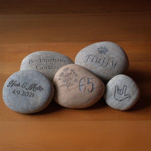 May include: Five engraved stones with different designs and text. One stone says "Fluffy" with a paw print, another says "Beckingham Cottage", and a third says "Nick & Molly 4.9.2021". The fourth stone has a floral design and the number "65". The last stone has a hand symbol.