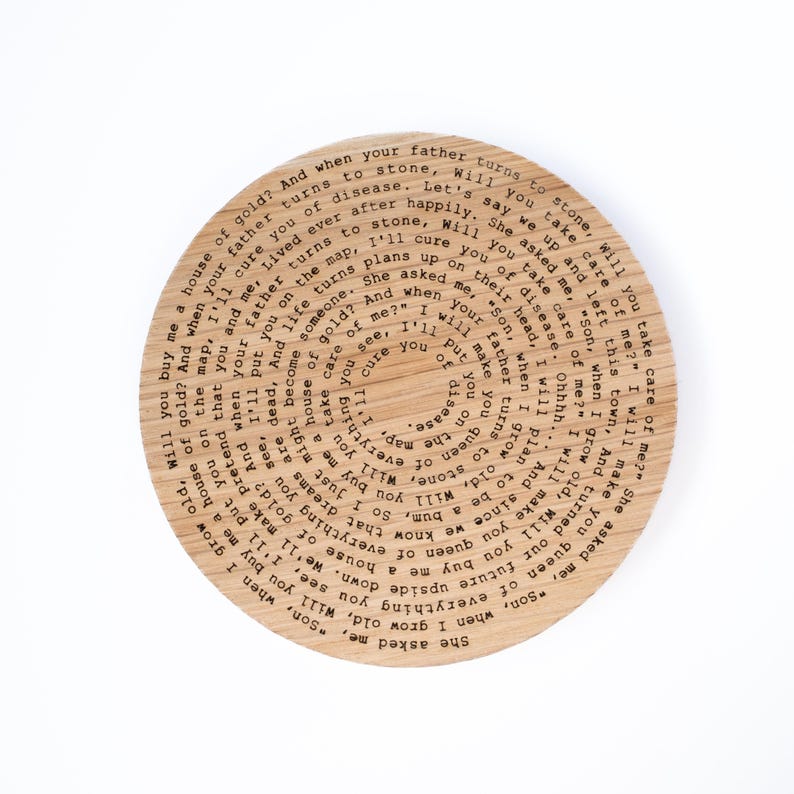 Puede incluir: Un posavasos redondo de madera con acabado marr&oacute;n claro. El posavasos presenta un dise&ntilde;o circular con texto grabado en la madera. El texto est&aacute; dispuesto en c&iacute;rculos conc&eacute;ntricos. Un art&iacute;culo decorativo.