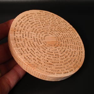 Puede incluir: Un posavasos redondo de madera con texto grabado. El posavasos est&aacute; hecho de madera de color claro y presenta un dise&ntilde;o circular con el texto en espiral hacia adentro. El texto es un poema o una cita. El posavasos es un art&iacute;culo decorativo.