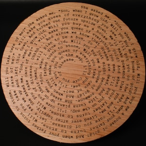 Puede incluir: Placa circular de madera con texto grabado con l&aacute;ser. El texto gira en espiral hacia adentro, con frases como "Will you take care of me?" y "Son, when I grow old". La placa es de madera clara y tiene una veta natural.