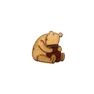 Peut inclure: Une broche en bois en forme d'ours assis tenant un pot de miel marron foncé. L'ours est de couleur beige clair avec des lignes gravées définissant ses oreilles, ses membres et ses traits. Le motif est composé de deux teintes de bois.