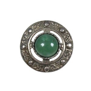 May include: A circular silver-toned brooch featuring an intricate embossed knotwork pattern along the outer rim, accented with four small raised spheres. A smooth, polished, round green stone is set securely in the center of the metal frame.