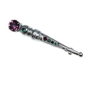 May include: Silver-toned brooch with a decorative design. The brooch features a large, round, purple gemstone at the top, followed by a smaller green gemstone. The body of the brooch is adorned with a series of smaller purple and green gemstones.