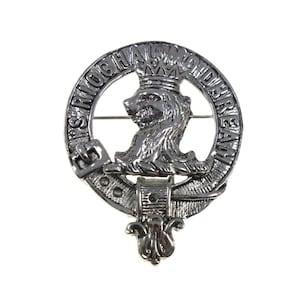 May include: Silver-toned metal brooch featuring a lion's head wearing a crown, encircled by text. The brooch has a pin on the back for attachment. The design is detailed and likely represents a family crest or emblem.