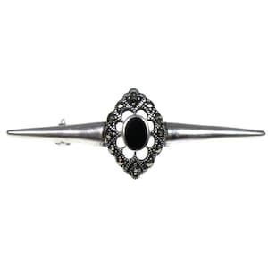 May include: A silver-toned brooch with a central oval black stone, surrounded by a decorative marcasite setting. The brooch has a bar shape with pointed ends, creating a symmetrical design. The jewelry piece is set against a white background.