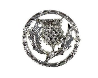 Puede incluir: Broche plateado con un diseño detallado de un cardo, rodeado por un borde decorativo. El cardo, símbolo de Escocia, es el foco central, con intrincados detalles en los pétalos y hojas. La broche se presenta sobre un fondo blanco.