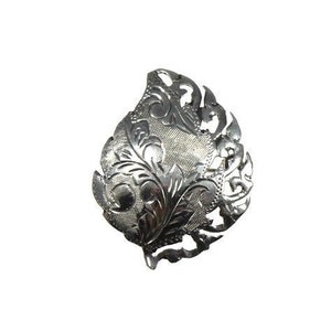 May include: A silver-colored, leaf-shaped brooch with intricate floral and scrollwork detailing. The brooch has a textured surface and a slightly curved shape, with a small loop at the bottom. The design is symmetrical and detailed.