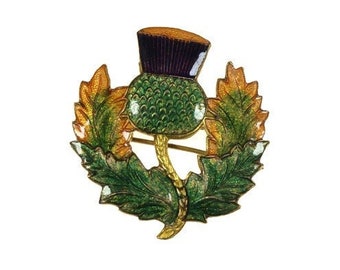 Puede incluir: Broche detallado con un diseño de cardo. El broche tiene una combinación de colores verde, dorado y morado. El cardo está rodeado de hojas verdes y doradas. El broche probablemente está hecho de metal y esmalte.