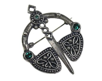 Puede incluir: Broche plateado con diseño de nudo celta. El broche presenta un diseño circular con un alfiler, dos elementos decorativos en forma de escudo y tres gemas verdes. El broche probablemente está hecho de metal.