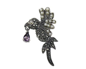May include: A silver-toned bird brooch with marcasite detailing and faux pearls. The bird holds a teardrop-shaped purple gemstone. The brooch has a detailed design and is set against a white background.