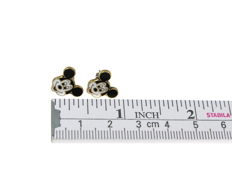 May include: A pair of gold-toned stud earrings featuring a Mickey Mouse design. The earrings have black and white enamel details. The ruler shows the earrings are approximately 0.5 inches in size.
