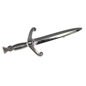 May include: A silver-toned metal brooch shaped like a miniature sword or dirk. The handle features a textured, ridged grip and a thistle-like pommel. The crossguard has curved ends and a small circular emblem at the center of the blade base.