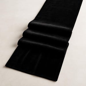 K&ouml;nnte beinhalten: Ein langer, rechteckiger Tischl&auml;ufer aus schwarzem Samt. Der L&auml;ufer ist mehrfach gefaltet, wodurch ein geschichteter Effekt entsteht. Der Stoff hat ein weiches, luxuri&ouml;ses Aussehen, das sich f&uuml;r formelle Abendessen oder Veranstaltungen eignet.