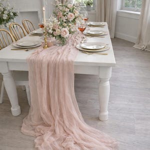 May include: A white dining table set for a special occasion. The table is adorned with a light pink, flowing table runner, floral centerpieces, and place settings. Gold-colored cutlery and candle holders add elegance.