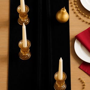 K&ouml;nnte beinhalten: Ein schwarzer Samt-Tischl&auml;ufer mit drei bernsteinfarbenen Glaskerzenhaltern, die jeweils eine wei&szlig;e Kerze halten. Eine goldene Birne und eine goldene Perlenkette liegen auf dem Holztisch. Ein wei&szlig;er Teller und eine rote Serviette sind ebenfalls zu sehen.